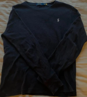 Mörkblå långärmad tröja Polo Ralph Lauren - Snygg mörkblå långärmad tröja från Polo Ralph Lauren i storlek M. Tröjan har ett diskret rutmönster i strukturen och klassisk rund hals. På bröstet sitter den ikoniska broderade loggan i vitt. Perfekt för dig som gillar stilrena och tidlösa plagg.