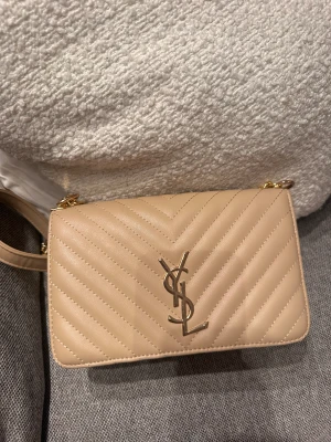 Beige quiltad axelväska från YSL - Snygg beige axelväska från YSL med guldiga detaljer och kedja. Väskan är quiltad med V-formade sömmar och har det klassiska YSL-emblemet framtill. Tillverkad i skinn och har en stilren, rektangulär form som ger en lyxig vibe.
