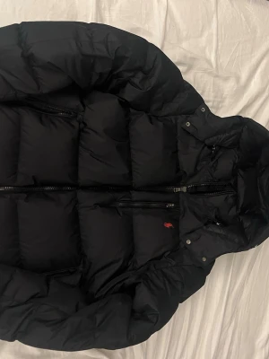 Svart dunjacka från Polo Ralph Lauren - Svart pufferjacka från Polo Ralph Lauren med röd broderad logga på bröstet. Jackan har huva, tvåvägsdragkedja och flera fickor med dragkedja. Perfekt för kalla dagar och har en klassisk, sportig look. Den är inpresipt aldrig använd pågrund av att den är för stor för mig. 