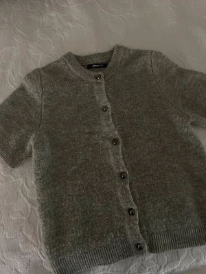 Grå kortärmad kofta från Gina Tricot - Mysig grå kofta från Gina Tricot i storlek S. Den har korta ärmar, rund hals och knäpps med silvriga knappar framtill. Materialet är stickat och känns mjukt mot huden, perfekt för lager på lager under höst och vår.