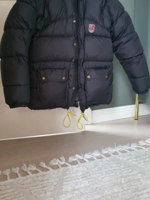 Svart dunjacka från Fjällräven - Svart pufferjacka från Fjällräven med klassisk logga på bröstet. Jackan har två stora fickor med knappar, gul snörning nedtill och dragkedja framtill. Otroligt varmt, perfekt för kalla dagar. Storlek XXXS men det passar XS-M då den är äldre modell.  Jag är 1.60, 44 kg och upplever den sitta för "loose" för min smak. 3 mycket små revor, par millimeter endast, se detaljbild. Priset går att diskutera (något)