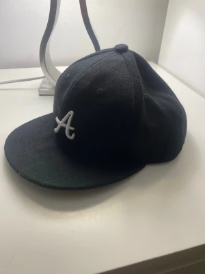 Svart keps med LA-patch och A-logga - Snygg svart keps med vit broderad A-logga framtill och en gul patch med texten 'Los Angeles AR 1985' på sidan. Klassisk form med böjd skärm och broderade detaljer. Perfekt för dig som gillar streetwear och sportig stil.