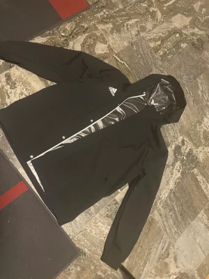 Svart regnjacka från Adidas med huva - Svart allvädersjacka från Adidas med vit logga på bröstet. Jackan har huva, långa ärmar och dragkedja framtill med tryckknappar. Tillverkad i syntetmaterial som står emot regn. Perfekt för blöta dagar och enkel att matcha med allt.