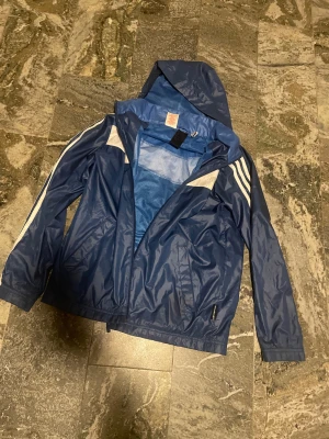 Blå Adidas vindjacka med huva - Snygg blå vindjacka från Adidas med klassiska vita ränder längs ärmarna och vit detalj över bröst och rygg. Jackan har huva, dragkedja framtill och meshfoder på insidan. Perfekt för blåsiga dagar och sportiga outfits. Storleken är junior 164. 