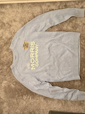 Ljusblå stickad tröja Morris Company - Snygg ljusblå stickad tröja från Morris Company i storlek M. Tröjan har ribbade muddar vid hals, ärmslut och nederkant samt ett broderat emblem och tryck framtill. Perfekt för dig som gillar klassisk stil med en modern touch. Det är i ett bra skick här användt endast några få gånger PM för mer frågor.😊