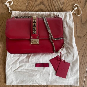 Röd axelväska från Valentino - Snygg röd axelväska från Valentino med kedjerem och coola guldfärgade nitar på framsidan. Väskan är tillverkad i skinn och har en stilren, rektangulär form. Perfekt för dig som vill ha en statement piece med lyxig vibe och detaljer som sticker ut.