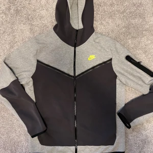 Nike grå och svart hoodie med dragkedja - Nike hoodie i grått och svart med hel dragkedja och huva. Snygg detalj med svart V-formad panel framtill och neongult Nike-logga på bröstet. Ficka med dragkedja på ena ärmen. Perfekt för chill eller träning.