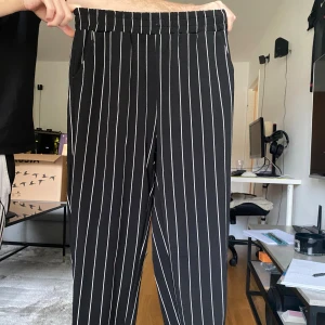 Svarta randiga byxor med resårmidja - Snygga svarta byxor med vita vertikala ränder och bekväm resår i midjan. Byxorna har raka ben och sidofickor, perfekta för en chill och stilren look. Passar dig som gillar streetstyle och vill ha något både bekvämt och trendigt.