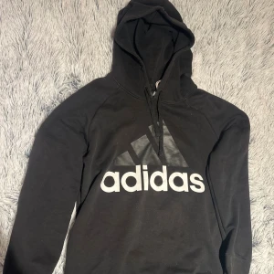 Svart Adidas hoodie - Svart hoodie från Adidas med stor vit logga framtill och mindre logga bak. Klassisk huva med snörning och långa ärmar. 