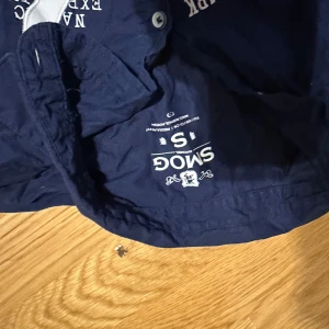 Mörkblå kortärmad skjorta SMOG S - Snygg mörkblå kortärmad skjorta från SMOG med vita tryck och siffran 45 på framsidan. Skjortan har knappar framtill och är gjord i mjuk bomull. Perfekt för dig som gillar sportig och avslappnad stil.