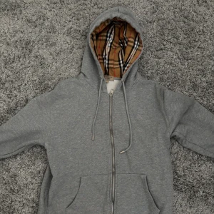 Grå Burberry hoodie med dragkedja - Säljer en grå hoodie från Burberry med klassiskt rutigt foder i huvan och snyggt broderat Burberry-märke på bröstet. Tröjan har dragkedja, snörning i huvan och två fickor framtill. Perfekt för dig som gillar stilren design med lyxig touch.