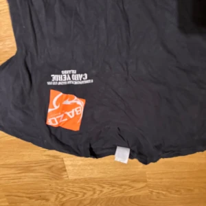 Svart t-shirt Cabo Verde Surf Society - Cool svart t-shirt från Bazor med stort tryck i vitt och orange på framsidan. Motivet har texten 'Cabo Verde Islands Surf Society' och en surfare i siluett. Perfekt för dig som gillar surf och streetstyle. Klassisk passform och rund hals.