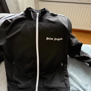 Palm Angels svart track jacket - Svart track jacket från Palm Angels med vita ränder längs ärmarna och vit dragkedja. Klassisk sportig look med hög krage och logga broderad på bröstet. Perfekt för dig som gillar streetwear och vill sticka ut med en ikonisk stil. Priset går att diskuteras vid snabb affär