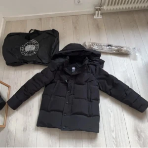Svart dunjacka från Canada Goose - Säljer en svart dunjacka från Canada Goose med klassisk patch på ärmen och avtagbar huva med beige pälsdetalj. Jackan har långa ärmar, knappar och dragkedja framtill. Perfekt för kalla vinterdagar och har en snygg, boxig passform.