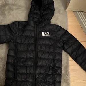 Svart dunjacka EA7 Emporio Armani - Mörkblå dunjacka från EA7 Emporio Armani med huva och dragkedja framtill. Jackan är quiltad och har en stilren logga på bröstet. Perfekt för kalla dagar och har en modern, sportig look. Använd ungefär 5 gånger