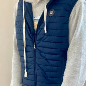 Mörkblå quiltad dunväst från Vinson - Snygg mörkblå dunväst från Vinson med quiltad design och dragkedja framtill. Västen har ett runt märke på bröstet med Vinson-logga och är perfekt att slänga över en hoodie. Klassisk passform och stilren look.