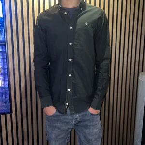 Mörkgrön skjorta från Morris - Snygg mörkgrön skjorta från Morris med klassisk button down-krage och vita knappar. Skjortan har broderad logga på bröstet och är tillverkad i bomull. Perfekt för dig som gillar stilrena och tidlösa plagg.