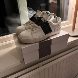 Valentino Rockstud sneakers vit/svart - Säljer ett par Valentino Rockstud sneakers i vitt skinn med svarta detaljer och ikoniska nitar på sidorna och runt hälen. Märket syns på sulan och tungan. Enda defekterna är att den är lite sliten på sulan och att en nit har lossnat uppe på tungan (Bild 5) annars är allt bra. Hör av er om ni har några frågor!☺️