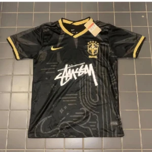Svart Brasil Nike x Stüssy fotbollströja - Kommer med dustpåse!!!!!!!!!!   Säljer en svart Brasil fotbollströja från Nike x Stüssy med gul krage och ärmslut. Tröjan har korta ärmar, Dri-Fit material och coolt grafiskt mönster på framsidan. Stüssy-loggan är tryckt i vitt och Brasil-emblemet i gult. Perfekt för dig som gillar streetwear och fotboll.
