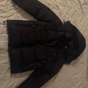 Svart pufferjacka Polo Ralph Lauren  - Svart pufferjacka från Polo Ralph Lauren med huva och dragkedja. Jackan har broderad röd logga på bröstet och flera praktiska fickor med dragkedja. Perfekt för kalla dagar och har en klassisk, sportig look. Helt i nyskick!