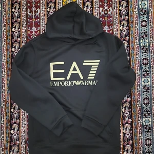 Svart hoodie EA7 Emporio Armani - Svart hoodie från EA7 Emporio Armani med stor logga i beige framtill. Klassisk modell med huva och känguruficka. Mjuk insida och långa ärmar, perfekt för en chill och stilren look. Den är slim-fit