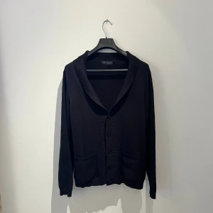 Svart kofta från Riley med ribbad krage - Svart kofta från Riley i storlek L med snygg ribbad krage och knappar framtill. Koftan har två praktiska fickor och är långärmad. Materialet är mjukt och känns som bomull, perfekt för lager-på-lager under höst och vinter. Hör av er vid frågor eller fler bilder