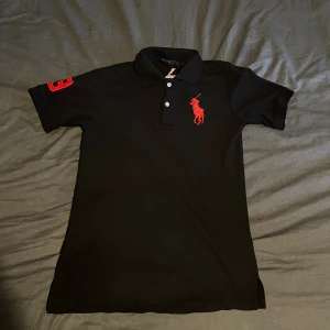 Svart Polo Ralph Lauren piké M - Svart pikétröja från Polo Ralph Lauren med röd broderad logga på bröstet och stort polospel-motiv på ryggen. Snygg röd siffra 3 på ärmen och broderad text på kragen. Klassisk krage och två knappar framtill. Tillverkad i 100% bomull. Börjar bli lite knopprig, hör av er vid frågor!