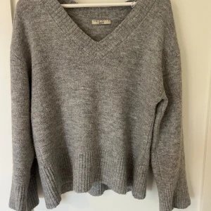 Grå stickad v-ringad tröja Gina Tricot - Mysig grå stickad tröja från Gina Tricot med v-ringning och breda ribbade muddar vid ärmslut och nederkant. Tröjan har en relaxed passform och är perfekt för kyliga dagar. Enkel att matcha med jeans eller kjol för en chill look.