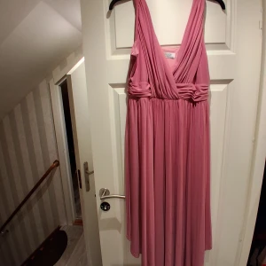 Lång rosa omlottklänning - Säljer en lång, rosa omlottklänning med djup v-ringning och breda axelband. Klänningen har fina draperingar i midjan och ett mjukt, fallande tyg som ger en elegant siluett. Perfekt för speciella tillfällen eller när du vill känna dig extra fin.