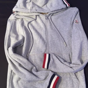Grå Moncler hoodie med ränder - Snygg grå hoodie från Moncler med dragkedja, huva och snörning. Ärmsluten och huvan har stilrena ränder i rött, vitt och marinblått. Klassisk logga på ärmen och mjukt bomullsmaterial som känns skönt mot huden.