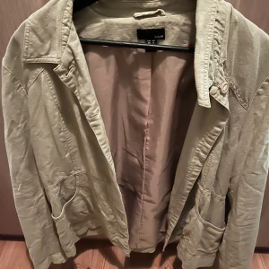 Beige kavajjacka från H&M - Stilren beige kavajjacka från H&M med klassisk krage, knappar och två stora fickor framtill. Jackan är tillverkad i ett mjukt bomullsmaterial och har en avslappnad passform som passar perfekt till många olika looks.