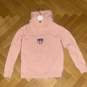 Ljusrosa hoodie från GANT - Säljer en ljusrosa hoodie från GANT med broderad logga och sköld på bröstet. Tröjan har huva, magficka och långa ärmar. Perfekt för dig som gillar klassisk och sportig stil.