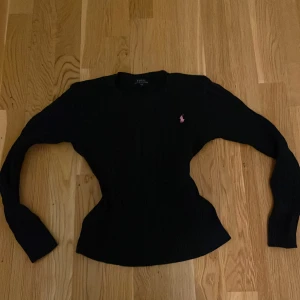 Mörkblå kabelstickad tröja Polo Ralph Lauren - Mörkblå kabelstickad tröja från Polo Ralph Lauren med klassisk rund hals och lång ärm. Tröjan har en rosa broderad logga på bröstet och är i mjuk bomull. Perfekt för dig som gillar stilrena och tidlösa plagg med en sportig touch.