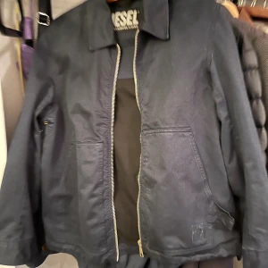 Svart vindjacka från Diesel - Snygg svart vindjacka från Diesel med klassisk krage och dragkedja framtill. Jackan har två stora fickor på sidorna och raka ärmar. Perfekt för dig som gillar stilren och enkel design. Den har lite fläckar som går bort om man visar den lite kärlek