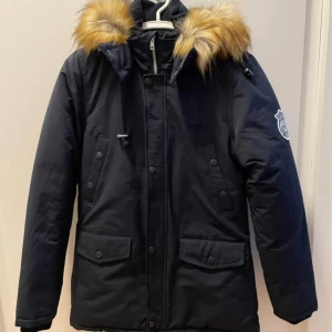 Mörkblå parkas från Svea med fuskpäls - Snygg mörkblå parkas från Svea med avtagbar huva och fuskpäls. Jackan har dragkedja och knappar framtill, stora fickor och Svea-logga på ärmen. Perfekt för kalla dagar med sin varma vaddering och klassiska look.