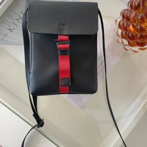 Svart axelväska Gaston Luga - NY  - Snygg svart axelväska från Gaston Luga med stilren design och röd detalj på framsidan. Väskan har ett lock med magnetstängning och justerbar axelrem. Materialet är slätt och vattenavvisande, perfekt för dig som gillar minimalistisk stil och vill ha med det viktigaste.