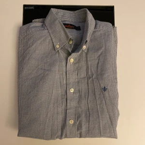 Rutig skjorta från Morris Stockholm - Snygg rutig skjorta från Morris Stockholm i storlek S. Skjortan är blå och vit med klassisk krage, långa ärmar och knappar framtill. Materialet är mjuk bomull och har ett broderat blått emblem på bröstet. Perfekt för dig som gillar stilrena detaljer.