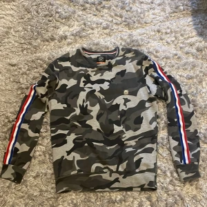 Camouflage sweatshirt med ränder - Snygg sweatshirt från Smith & Jones med grått, svart och mörkgrönt camouflage-mönster. Långärmad modell med coola röda, vita och blå ränder längs ärmarna. Rund hals och mjukt bomullsmaterial, perfekt för en streetwear-look.