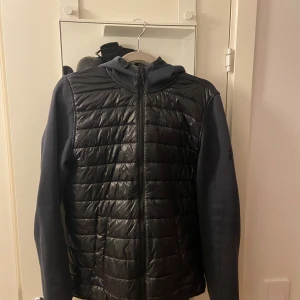 Svart hybridjacka Peak Performance S - Snygg svart hybridjacka från Peak Performance i storlek S. Jackan har quiltad framsida i glansigt material, mjuka ärmar och rygg samt huva. Dragkedja framtill och sportig look, perfekt för kyliga dagar.