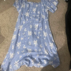 Blå blommig klänning från 157 - Supersöt ljusblå klänning från 157 med vita blommor, kort ärm och smockad rygg. Klänningen har en lätt utställd form och knappar framtill. Perfekt för sommarens alla tillfällen!