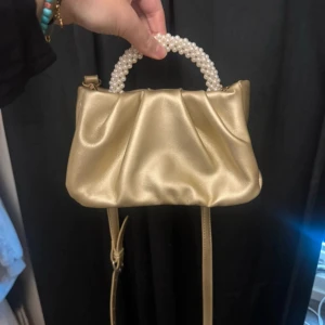 Guldig handväska med pärlhandtag - Snygg guldig handväska med glansigt material och ett handtag dekorerat med pärlor. Väskan har en mjuk, rynkad form och magnetlås. Perfekt storlek för mobil och småsaker, med avtagbar axelrem för olika stilar.