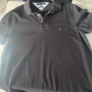 Svart pikétröja Tommy Hilfiger XL - Klassisk svart pikétröja från Tommy Hilfiger i storlek XL. Tröjan har korta ärmar, krage med knappar och den ikoniska lilla Hilfiger-loggan broderad på bröstet. Tillverkad i bomull för en skön och luftig känsla. Oanvänd 10/10 skick!