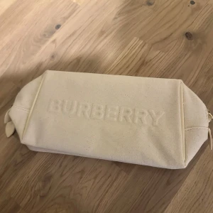 Beige Burberry väska i bomull taske - Snygg beige kuvertväska från Burberry i bomull med präglad logga på framsidan. Väskan har två dragkedjor med guldfärgade detaljer och praktisk innerficka. Perfekt för att hålla koll på dina essentials och ge din look en clean vibe.