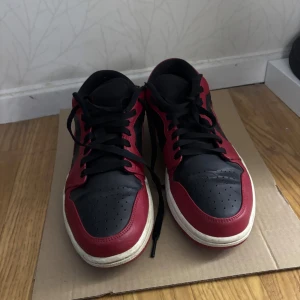 Nike Air Jordan 1 Low svart/röd - Nike Air Jordan 1 Low sneakers i svart och rött med klassisk Swoosh på sidan och Air Jordan-logga på hälen. Skorna har snörning, rund tå och platt sula. Tillverkade i skinn med perforerade detaljer på tån för extra ventilation. Perfekt för dig som gillar streetwear och ikoniska sneakers.