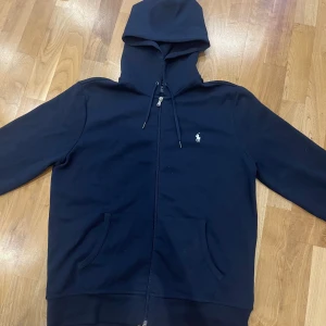 Marinblå hoodie från Polo Ralph Lauren - Säljer en marinblå hoodie från Polo Ralph Lauren med dragkedja och huva. Tröjan har två fickor framtill, snörning i huvan och den klassiska vita loggan broderad på bröstet. Storlek M, har använt den max 5 gånger riktigt bra skick.  köpte den från polo Ralph laurens hemsida. 