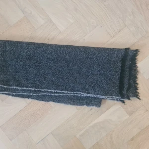 Mörkgrå stickad halsduk med fransar - En lång och mysig mörkgrå halsduk med diskret melerat mönster och fransar i ändarna.