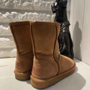 Klassiska bruna UGG boots i mocka - Säljer ett par klassiska UGG boots i ljusbrun mocka med rund tå och platt sula. Insidan är fodrad med mjuk päls för extra värme. Perfekta för kyliga dagar och har den ikoniska UGG-loggan bak på hälen. ALDRIG ANVÄNDA 