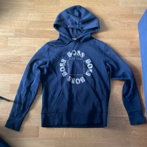 Svart hoodie från Hugo Boss - Snygg svart hoodie från Hugo Boss med stort BOSS-tryck i guld och vitt framtill. Tröjan har huva med dragsko, långa ärmar och en klassisk känguruficka. Perfekt för en chill och stilren look. Den har lite färg på sig och är lite sol bränd storlek xs män passar s, pris kan diskuteras 