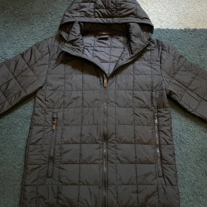 Svart quiltad pufferjacka från Everest - Svart pufferjacka från Everest med quiltad design och huva. Jackan har två sidofickor med dragkedja och snyggt Everest-märke nedtill. Perfekt för kalla dagar och har en stilren look som funkar till det mesta. Köpte den precis men jag gillade inte den riktigt.