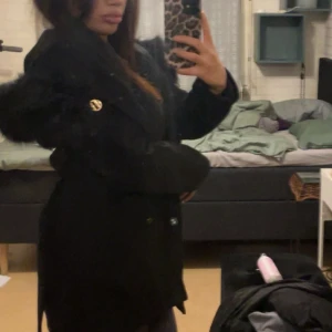 Svart kappa Hollies - Snygg svart kappa från Hollies med fuskpäls runt huvan och knytskärp i midjan. Kappan har en klassisk siluett, långa ärmar och stängs med knappar framtill. Perfekt för dig som vill ha en stilren och varm jacka med lyxig känsla.